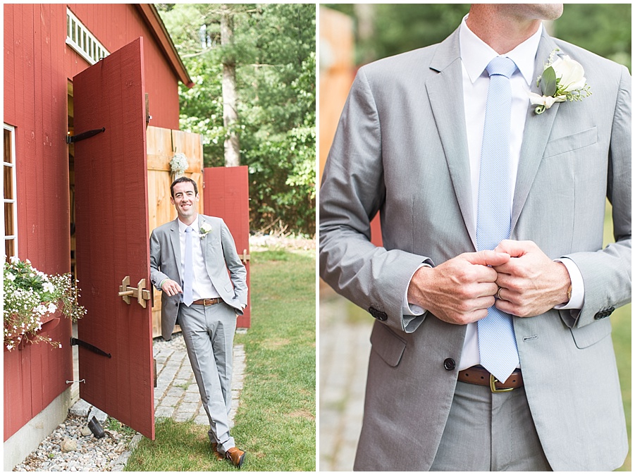 groom stands in his grey suit in front of red barn 